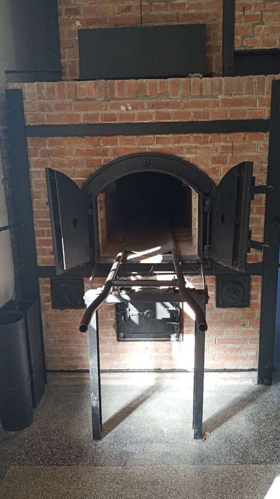 Een afbeelding van een van de crematoria in voormalig kamp Vught. In een gemetselde muur zitten zwarte deuren van staal, deze staan naar buiten open, met aan de voorkant een soort stalen brancard om de overledene in de oven te kunnen schuiven.