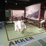 Voor op de grond staat "Akker", met daar een witte koe op en verschillende landwerktuigen vanuit de 18e eeuw erachter.