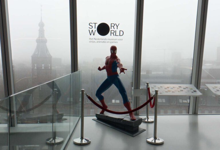 Een pop van spiderman staat voor een glaswand met daarachter een logo van Story World