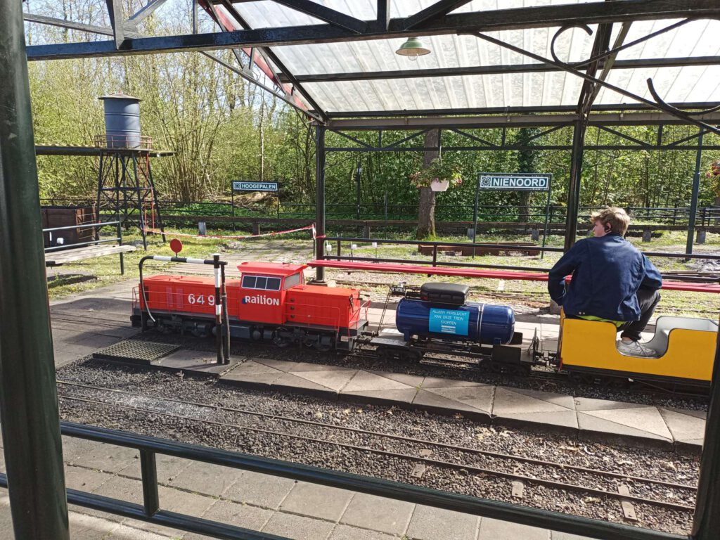 Een miniatuur rode dieseltrein met daarachter een blauw persluchtvat. Daarachter een eerste wagon welke geel is en waar iemand in zit.