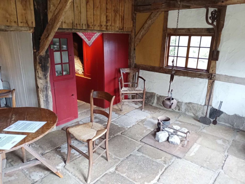 Op de voorgrond een sobere tafel van hout en een stoel. Een plek voor vuur maken en een bedstee in het rood.