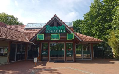 Op de foto staat de entree van Tierpark Nordhorn. Dit is vorm gegeven in een soort huisje met glazen schuifdeuren en in het groen de naam Tierpark Nordhorn aan de voorkant.